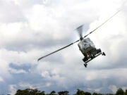 Gray military helicopter in flight above treeline under a cloudy sky, rotor blades blurred.