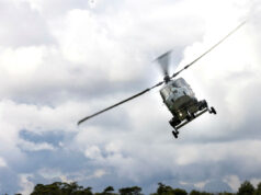 Thales wins deal to run Britain’s Link 16 network Gray military helicopter in flight above treeline under a cloudy sky, rotor blades blurred.