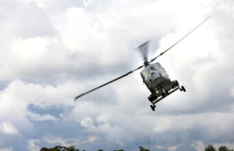 Gray military helicopter in flight above treeline under a cloudy sky, rotor blades blurred.