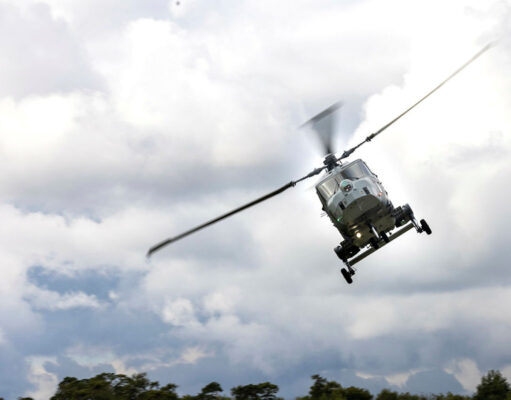Gray military helicopter in flight above treeline under a cloudy sky, rotor blades blurred.