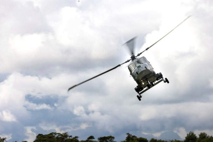 Gray military helicopter in flight above treeline under a cloudy sky, rotor blades blurred.