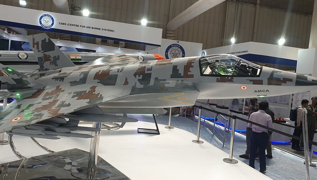 Side view of a camouflaged fighter jet on display at an air show exhibit, with cockpit visible and visitors nearby.