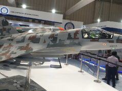 Side view of a camouflaged fighter jet on display at an air show exhibit, with cockpit visible and visitors nearby.