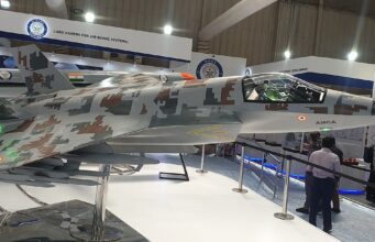 Side view of a camouflaged fighter jet on display at an air show exhibit, with cockpit visible and visitors nearby.