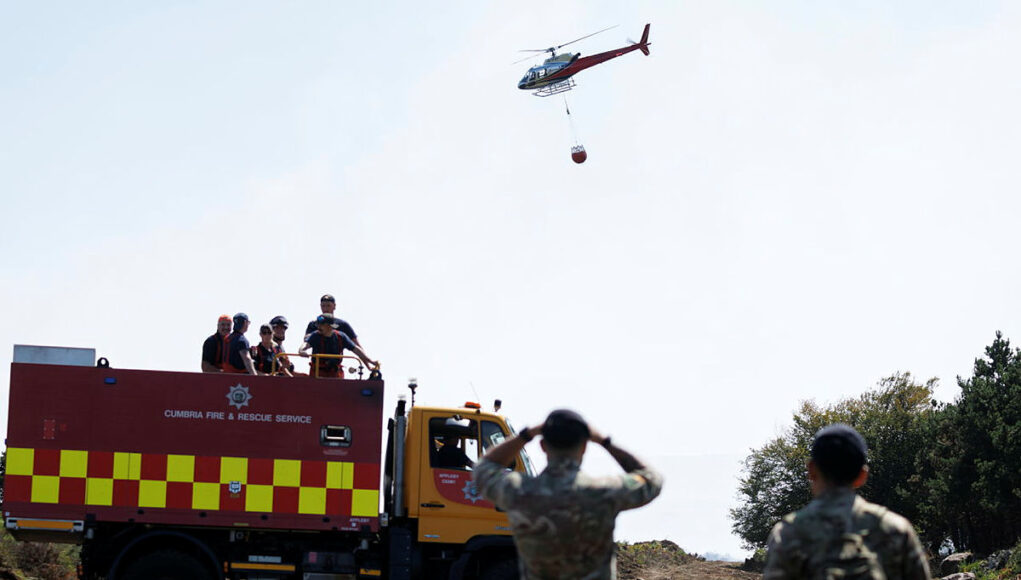 ARMY039-OFFICIAL-20260815-077.jpg.iCfoudWEMtFDiwllYBMA.Hw5yZpiJOR (1) Fire and rescue truck with crew on top, helicopter overhead delivering a water bucket in a rural area, soldiers observing in the foreground.