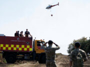 Army deploys 200 troops to tackle Welsh wildfires Fire and rescue truck with crew on top, helicopter overhead delivering a water bucket in a rural area, soldiers observing in the foreground.