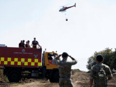 Army deploys 200 troops to tackle Welsh wildfires Fire and rescue truck with crew on top, helicopter overhead delivering a water bucket in a rural area, soldiers observing in the foreground.