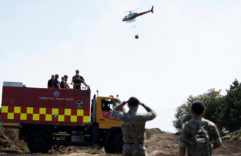 Army deploys 200 troops to tackle Welsh wildfires Fire and rescue truck with crew on top, helicopter overhead delivering a water bucket in a rural area, soldiers observing in the foreground.