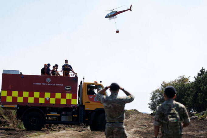 Fire and rescue truck with crew on top, helicopter overhead delivering a water bucket in a rural area, soldiers observing in the foreground.