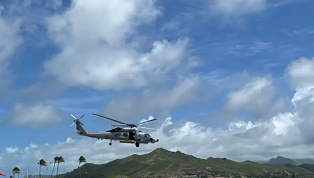 Gray military helicopter hovering low above an airstrip by the coast, with green hills and blue sky in the background.