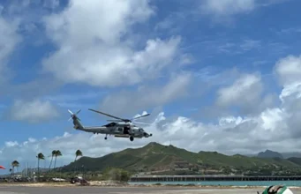 AI submarine hunter flown on American helicopters Gray military helicopter hovering low above an airstrip by the coast, with green hills and blue sky in the background.