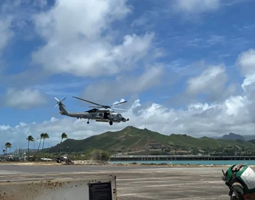 AI submarine hunter flown on American helicopters Gray military helicopter hovering low above an airstrip by the coast, with green hills and blue sky in the background.