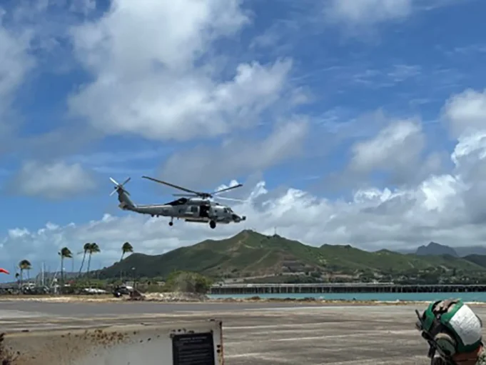AI submarine hunter flown on American helicopters Gray military helicopter hovering low above an airstrip by the coast, with green hills and blue sky in the background.
