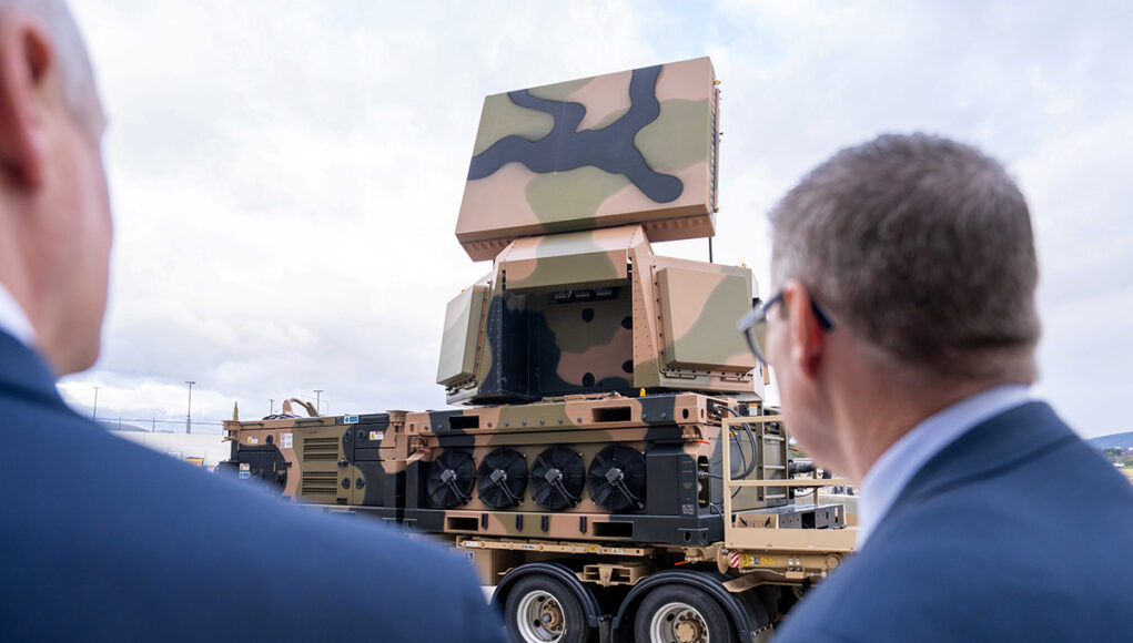 CEA1 Two men in suits watch a camouflaged mobile artillery/launcher mounted on a large wheeled transport truck outdoors