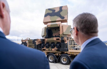 Two men in suits watch a camouflaged mobile artillery/launcher mounted on a large wheeled transport truck outdoors