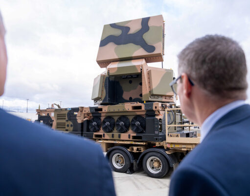 Two men in suits watch a camouflaged mobile artillery/launcher mounted on a large wheeled transport truck outdoors