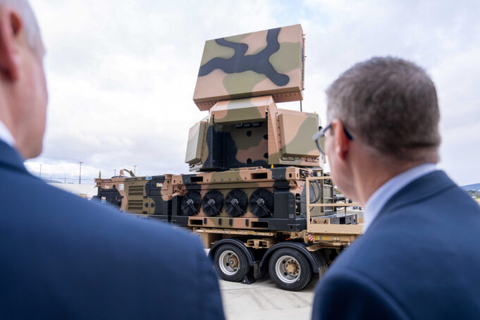 Two men in suits watch a camouflaged mobile artillery/launcher mounted on a large wheeled transport truck outdoors