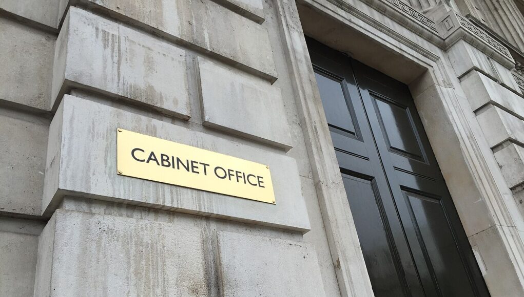 Cabinet_Office_(19243836200) Gold plaque reading 'CABINET OFFICE' mounted on a weathered stone wall beside a tall black door with brass handles.