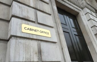 Gold plaque reading 'CABINET OFFICE' mounted on a weathered stone wall beside a tall black door with brass handles.