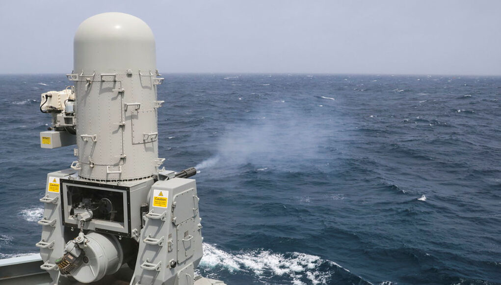 Naval gun turret on a gray battleship or naval vessel, facing the open ocean with waves in the background
