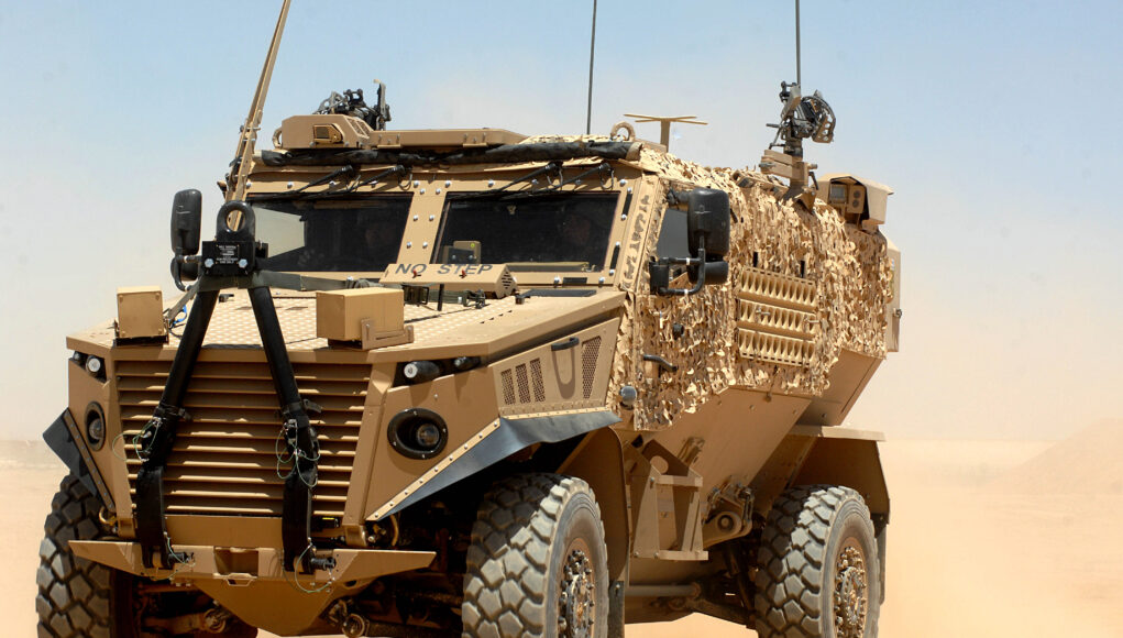 Foxhound Patrol Vehicle in Afghanistan Desert armored military vehicle in tan camouflage on sandy terrain under a clear blue sky, front view showing heavy tires and equipment on the hood and sides.