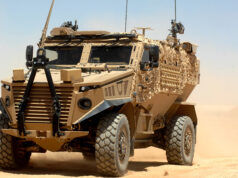 Hybrid Foxhound and Boxer could carry lasers, paper says Desert armored military vehicle in tan camouflage on sandy terrain under a clear blue sky, front view showing heavy tires and equipment on the hood and sides.