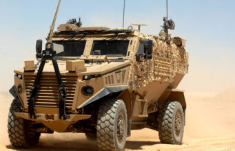 Hybrid Foxhound and Boxer could carry lasers, paper says Desert armored military vehicle in tan camouflage on sandy terrain under a clear blue sky, front view showing heavy tires and equipment on the hood and sides.