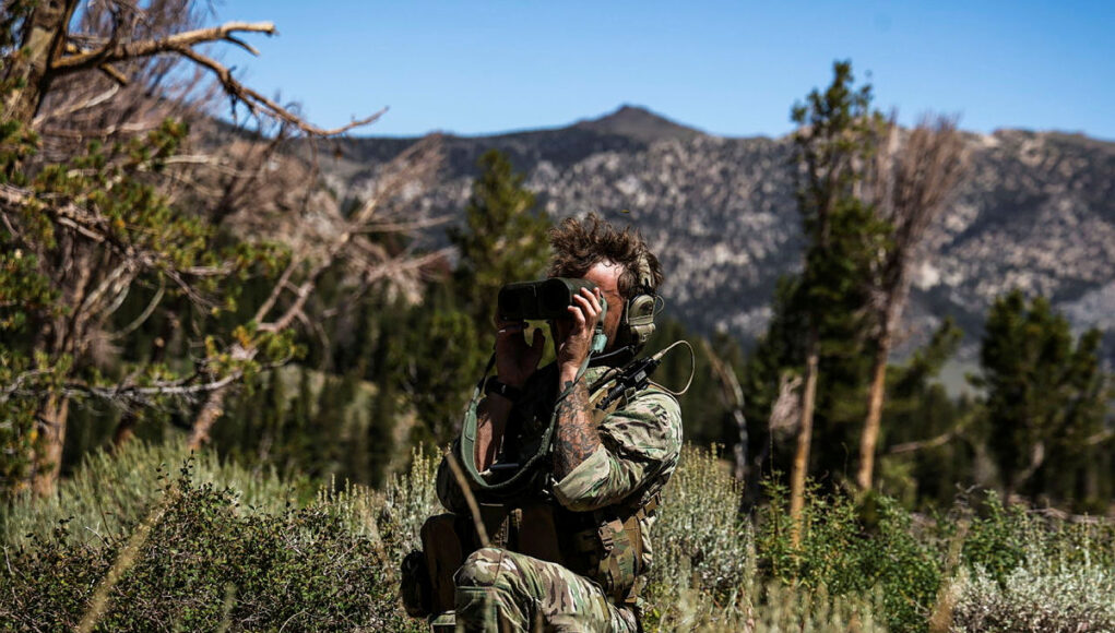 FLEET-20250817-XC0158-276.jpg.iCeah7iEMhFtR-1jYBMA.ZM2DY_pTHq Soldier in camouflage kneels in tall grass, using binoculars to scout distant mountains.