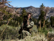 Soldier in camouflage kneels in tall grass, using binoculars to scout distant mountains.