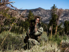 Soldier in camouflage kneels in tall grass, using binoculars to scout distant mountains.