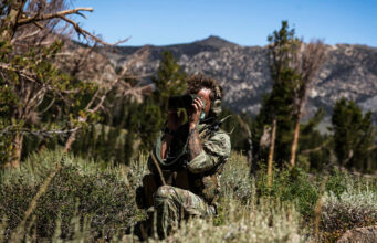 Soldier in camouflage kneels in tall grass, using binoculars to scout distant mountains.