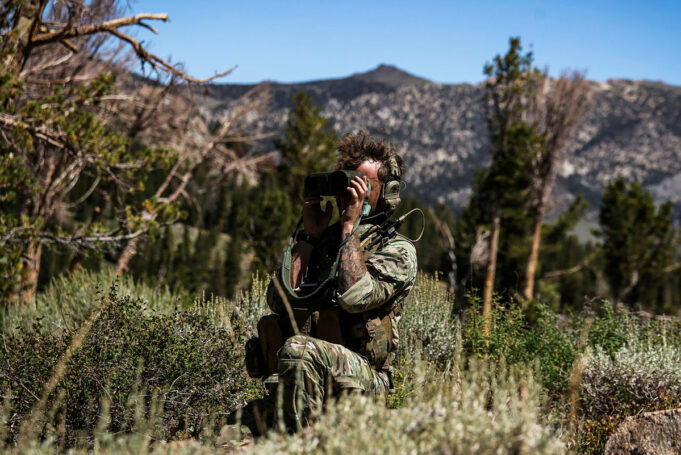 Thermal cameras that soldiers could carry are in view Soldier in camouflage kneels in tall grass, using binoculars to scout distant mountains.