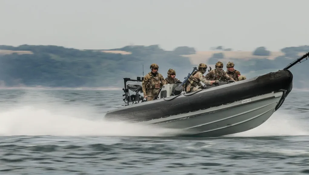 FLEET-20260228-ZI0081-022 Six soldiers in camouflage gear on a high-speed inflatable military boat skimming over the water with a misty shoreline in the background.