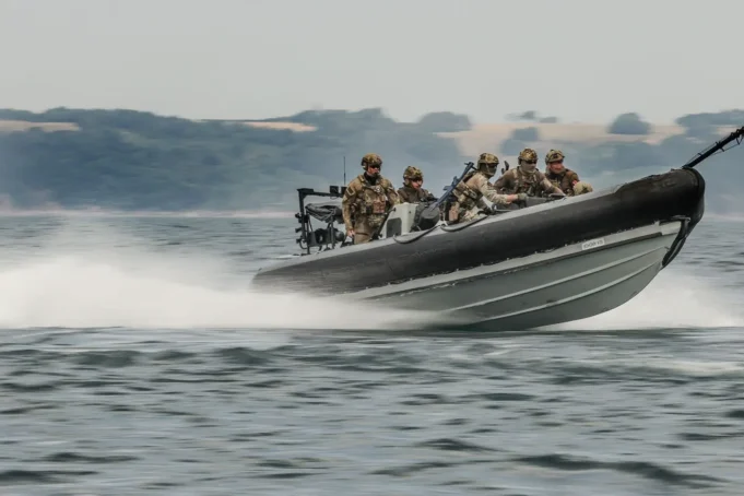 Six soldiers in camouflage gear on a high-speed inflatable military boat skimming over the water with a misty shoreline in the background.