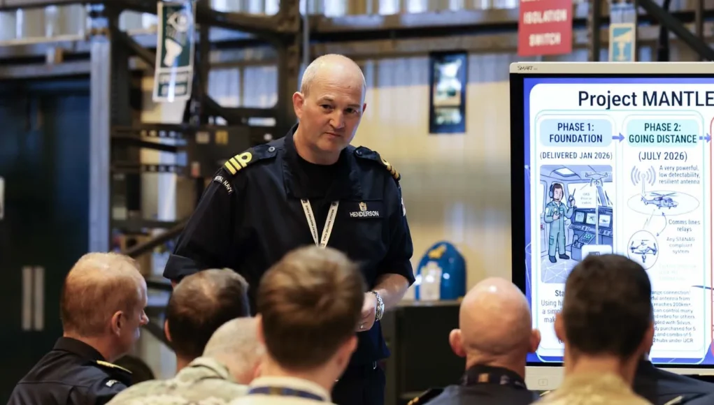 FLEET-20260727-XH0058-007 Military officer addresses a seated audience beside a large Project Mantle infographic board in a briefing room/hangar.