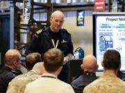 Royal Navy tests long-range drone control network Military officer addresses a seated audience beside a large Project Mantle infographic board in a briefing room/hangar.