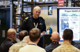 Military officer addresses a seated audience beside a large Project Mantle infographic board in a briefing room/hangar.