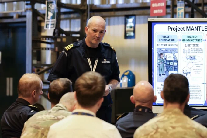 Royal Navy tests long-range drone control network Military officer addresses a seated audience beside a large Project Mantle infographic board in a briefing room/hangar.