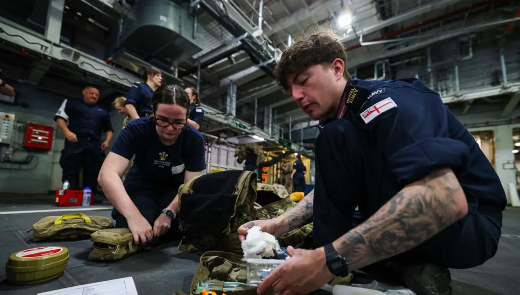 FRPUE-20260709-XF0123-007 British Royal Navy personnel kneel on a hangar deck, organizing and packing medical supplies from tactical bags for a field drill or mission.