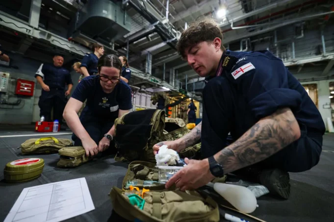 First combat medic course run aboard a Royal Navy ship British Royal Navy personnel kneel on a hangar deck, organizing and packing medical supplies from tactical bags for a field drill or mission.