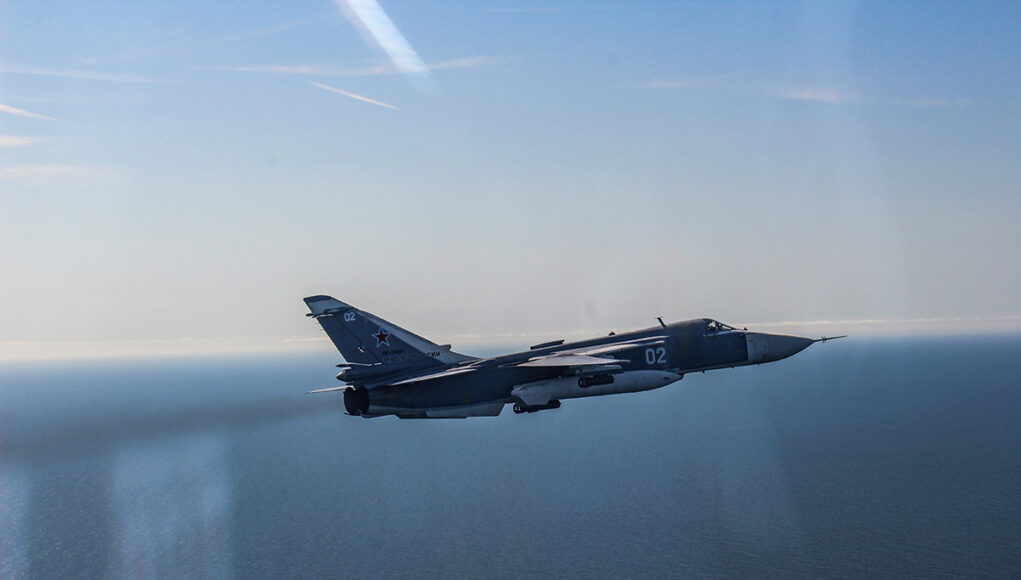 HO4HkjrWQAAbiAw Blue military fighter jet with the number 02 flying over the calm ocean against a clear blue sky, navy-gray livery visible on the fuselage and tail insignia clearly shown.