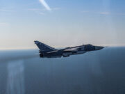 Blue military fighter jet with the number 02 flying over the calm ocean against a clear blue sky, navy-gray livery visible on the fuselage and tail insignia clearly shown.