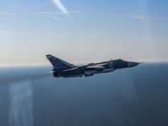 Blue military fighter jet with the number 02 flying over the calm ocean against a clear blue sky, navy-gray livery visible on the fuselage and tail insignia clearly shown.