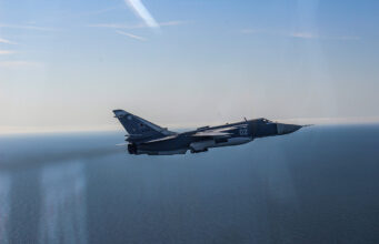 Blue military fighter jet with the number 02 flying over the calm ocean against a clear blue sky, navy-gray livery visible on the fuselage and tail insignia clearly shown.