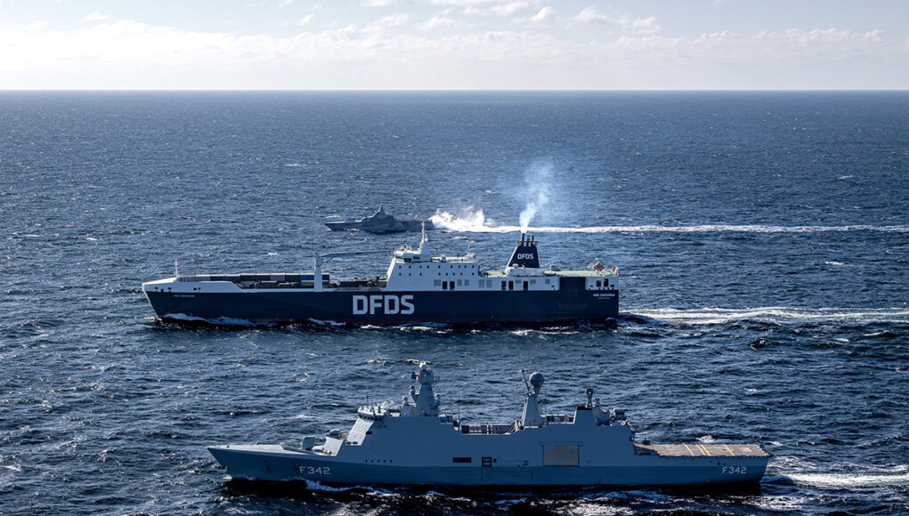 HPgvoQUXEAAw1zG Two large ships sail on open sea, a blue DFDS ferry behind a gray military vessel labeled F342.