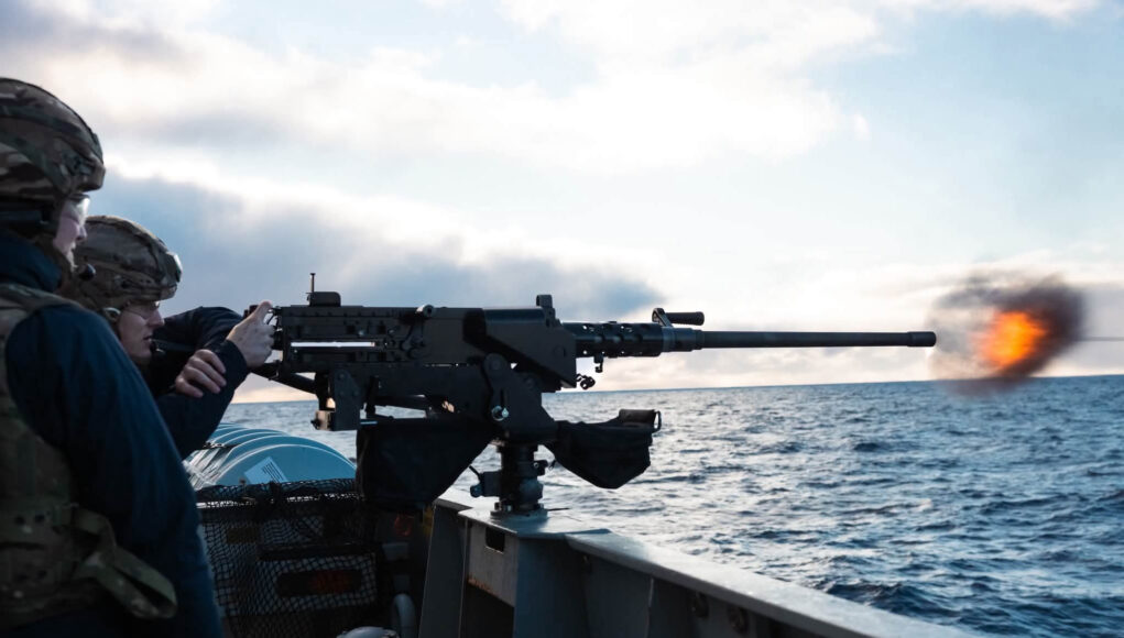 HPiYYgWXMAASdqK Sailors on a naval deck firing a large mounted gun, muzzle flash over the ocean with a cloudy sky behind.