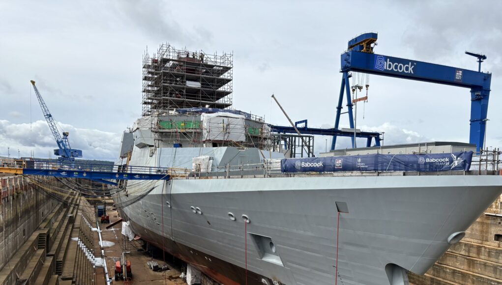 Dry docked warship under heavy scaffolding and construction cranes, with a blue Babcock crane overhead and workers on the upper level.