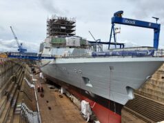 Dry docked warship under heavy scaffolding and construction cranes, with a blue Babcock crane overhead and workers on the upper level.