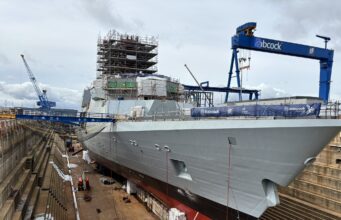 Multiple new British frigates taking shape in Rosyth Dry docked warship under heavy scaffolding and construction cranes, with a blue Babcock crane overhead and workers on the upper level.