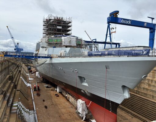 Multiple new British frigates taking shape in Rosyth Dry docked warship under heavy scaffolding and construction cranes, with a blue Babcock crane overhead and workers on the upper level.